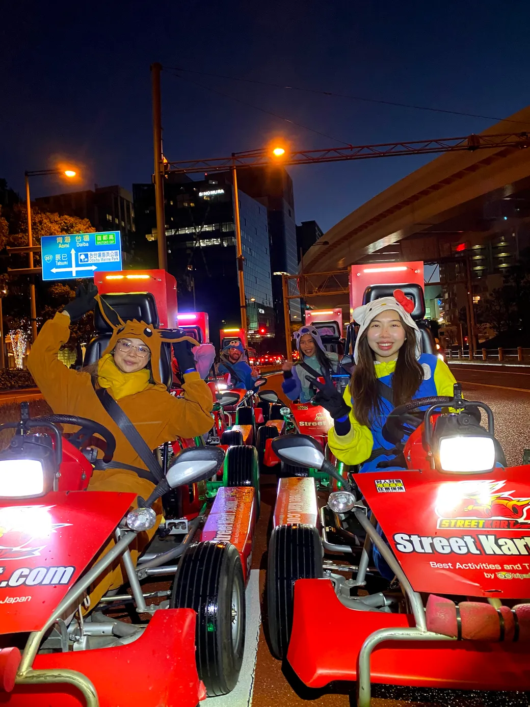 People in colorful animals costumes ride red go-karts on a city street at night, smiling and posing for the camera.