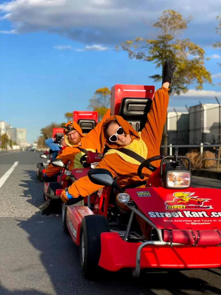 IMG_3604 - StreetKart Group of people in orange costumes riding red go-karts down a city street, foreground driver raising an arm in the air.