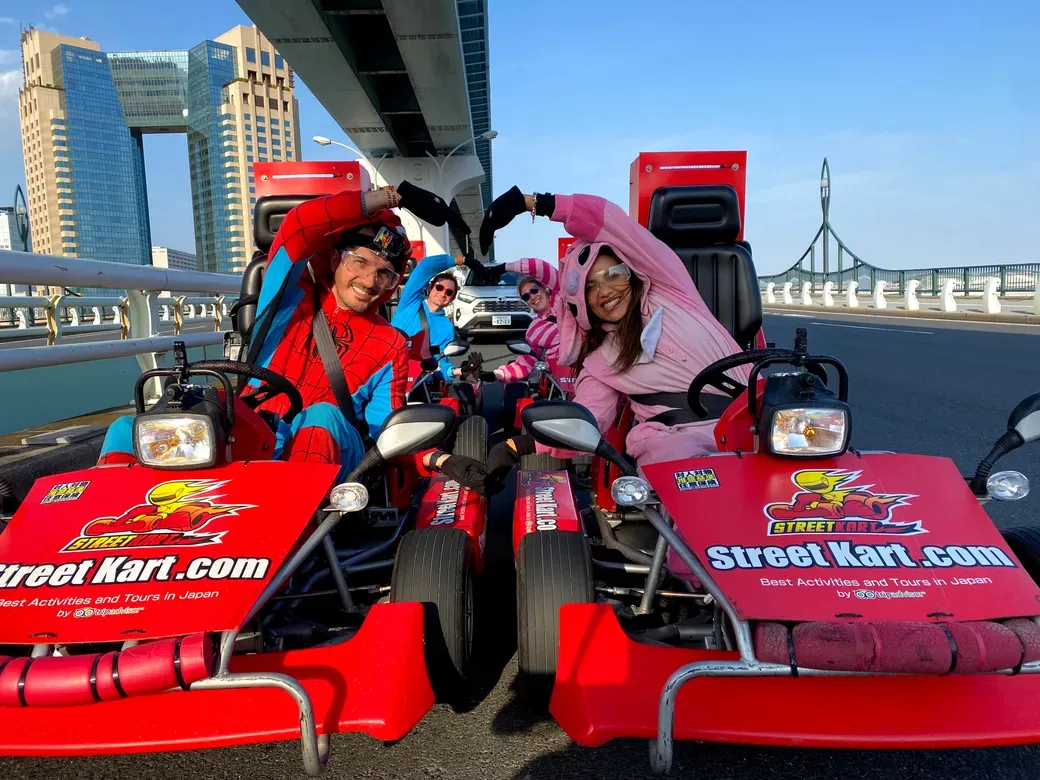 Four people in colorful costumes sit in red Street Kart go-karts on a sunny city bridge, making a heart shape with their arms.